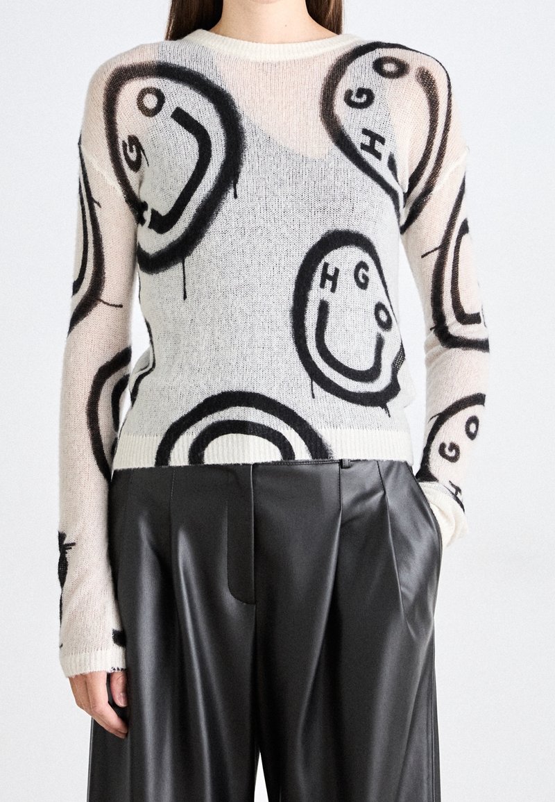 Pull beige clair avec des visages souriants abstraits noirs et des lettres, associé à un pantalon large en cuir noir taille haute.