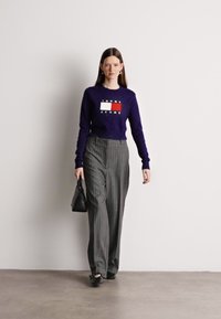 Μωβ πουλόβερ με λογότυπο στο στήθος, συνδυασμένο με γκρι ριγέ παντελόνι ψηλής μέσης. Το μοντέλο κρατάει μια μικρή μαύρη τσάντα και φοράει μαύρα παπούτσια.