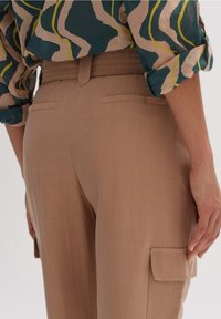 Béžové cargo kalhoty s hladkou texturou, švy a dvěma zadními kapsami. Pás má jemné sklady pro vylepšení designu.