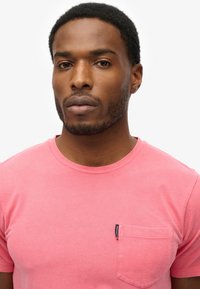 Camiseta de algodón de manga corta y cuello redondo en un vibrante color rosa, con un bolsillo en el pecho. Etiqueta visible en el bolsillo, textura suave.