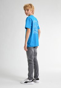 Camiseta de algodón turquesa con un gráfico floral en la parte posterior, combinada con jeans grises y zapatillas blancas con detalles en negro.