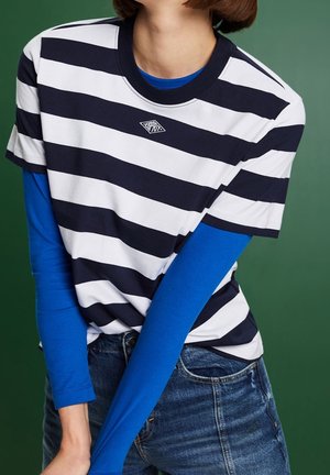 Persoon die een zwart-wit horizontaal gestreept shirt met korte mouwen draagt over een felblauw shirt met lange mouwen en een blauwe spijkerbroek, met een groene achtergrond.