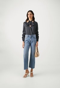 Blusa blu navy con pois e dettagli a fiocco, jeans azzurri a vita alta, borsa beige e sandali con tacco color crema, caratterizzati da un orlo frangiato.