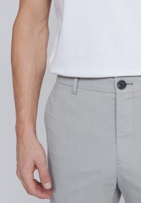 Pantaloni gri, texturat, cu închidere cu nasture și curea, asortați cu o cămașă albă cu mânecă scurtă. Mână poziționată pe țesătură.