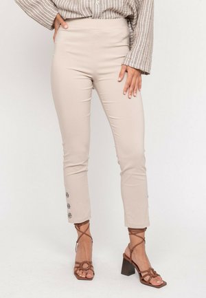 Femme portant un pantalon beige taille haute et slim avec des détails de boutons près des chevilles, accompagnée de sandales à talons carrés brun à brides, sur fond blanc.