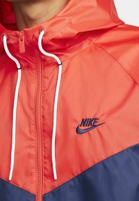 Nike windbreaker featuring a bright coral top and navy bottom, with white drawstrings and a navy logo on the chest. Lightweight, smooth texture.