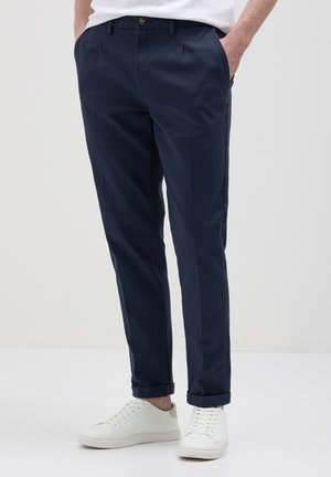 Uomo che indossa pantaloni slim-fit blu navy con risvolti e sneakers bianche, con le mani in tasca, in piedi su uno sfondo chiaro e semplice.