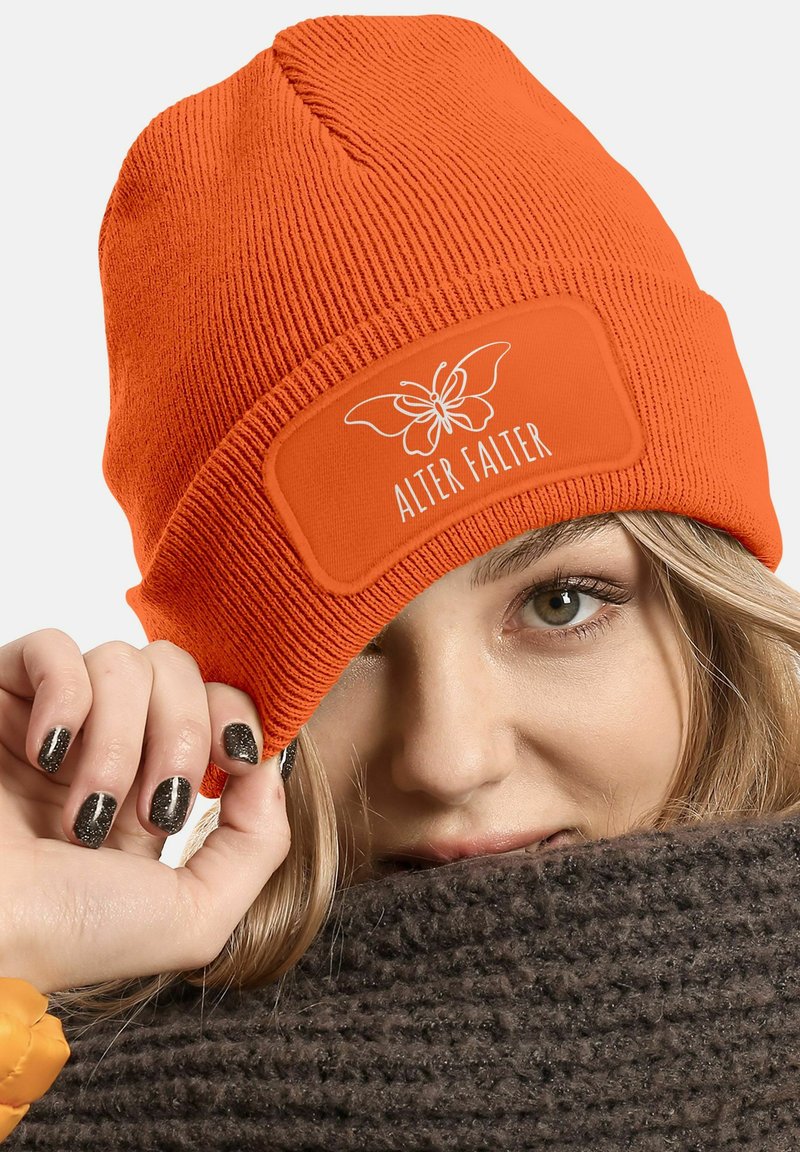 Orange knit beanie with a flat label featuring a butterfly graphic and “ALTER FALTER” text, paired with a chunky brown scarf.