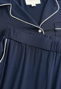 Marineblaues Pyjama-Set aus glattem Stoff; besteht aus einem Oberteil mit Kragen, weißer Paspelierung und einem elastischen Bund mit Knopfdetail am Oberteil.