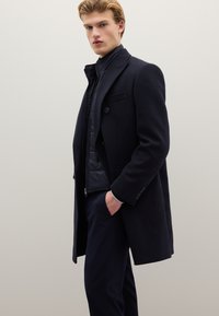 Cappotto in lana blu navy con vestibilità sartoriale, caratterizzato da un davanti doppiopetto, colletto a lancia e uno strato interno trapuntato.