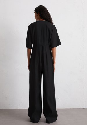 Combinaison noire avec des pantalons à jambes larges, des manches courtes et une taille cintrée. Fabriquée dans un tissu lisse, avec un design épuré et minimaliste.