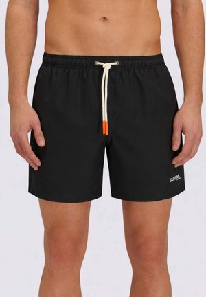 Uomo che indossa pantaloncini da bagno neri con estremità dei laccetti bianche e logo "SUNDEK" sulla gamba sinistra, in piedi su uno sfondo semplice.