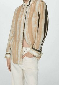 Persona che indossa una camicia a righe beige e bianca, con bottoni e maniche lunghe, insieme a pantaloni a vita alta di colore chiaro, con una mano in tasca.