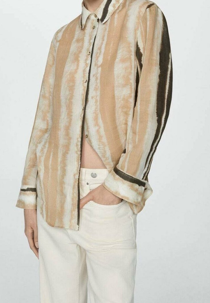 Persona che indossa una camicia a righe beige e bianca, con bottoni e maniche lunghe, insieme a pantaloni a vita alta di colore chiaro, con una mano in tasca.
