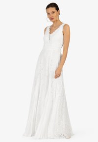 Robe en dentelle blanche avec un corsage ajusté, décolleté en V profond et bords festonnés. Jupe fluide avec une texture subtile et des motifs floraux. Design élégant.