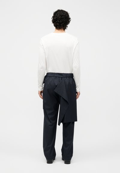 Άνδρας με κατσαρά μαλλιά που φοράει λευκό πουκάμισο μακριάς μανίκας και μαύρο παντελόνι με ρίγες, με ασύμμετρη υφασμάτινη λεπτομέρεια που κρέμεται στο πίσω μέρος.