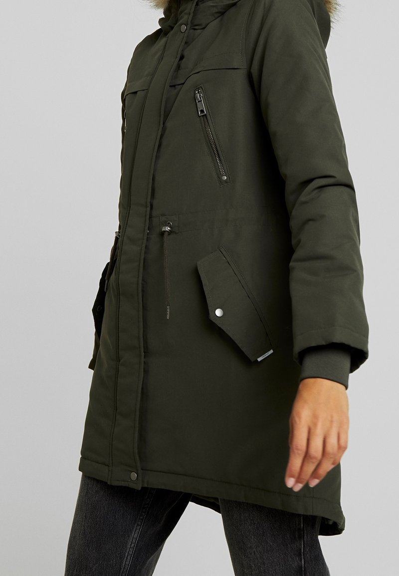 Grön vadderad parka-jacka med huva fodrad med päls, dragkedjeförsedda bröst- och sidofickor samt kontrasterande ärmslut. Slät textur med matt yta.