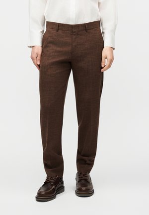 Man wearing brown textured slim-fit trousers, dark brown leather lace-up shoes, and a white long-sleeve button-up shirt, standing.