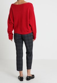 Röd överdimensionerad tröja med bred halsringning, i kombination med grå rutiga byxor. Svarta platta skor kompletterar outfiten. Mjuk textur synlig.