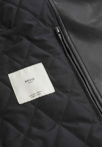 Tessuto nero trapuntato con un dettaglio in pelle liscia; presenta un'etichetta bianca con il nome del marchio e le istruzioni per la cura. La cerniera è visibile.