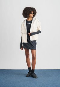 Γυναίκα με σγουρά μαλλιά που φοράει γυαλιά ηλίου, κρεμ φούτερ με φερμουάρ, μαύρο μακρυμάνικο τοπ, μαύρη φούστα, μαύρες κάλτσες και μαύρα αθλητικά παπούτσια στέκεται σε μπλε μοκέτα.