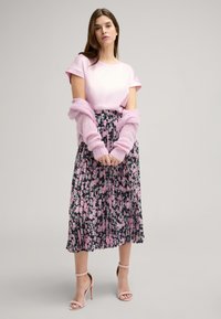 Lichtroze top met korte mouwen, gecombineerd met een geplooide, zwart bloemenrok met roze en groene accenten. Roze gebreide sjaal en hakken met bandjes.