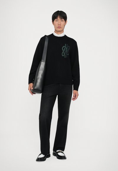 Young man wearing a black sweater with a green design, black pants, white shirt, black loafers with gold detail, and a black shoulder bag.