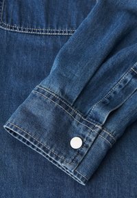 Dettaglio di una camicia di denim con tessuto blu, cuciture visibili e polsini arrotondati con un accentuato bottone argento.