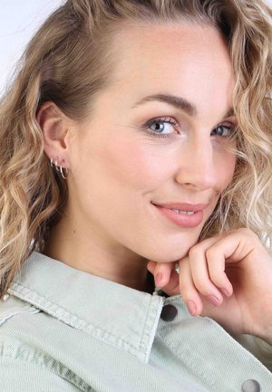 Zilveren hoepeloorbellen met drie verstrengelde ringen; model draagt een lichtgroene denimjas met zichtbare stiksels en knoopdetails.