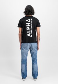 T-shirt preta de algodão com grande texto branco "ALPHA INDUSTRIES" nas costas. Combinada com jeans de denim azul claro e ténis pretos.