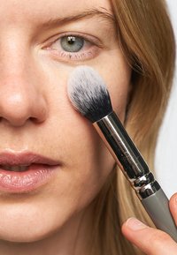 Makeupborste med grått handtag och silverfäste, som applicerar produkt på kinden hos en person med blont hår och ljus hy.