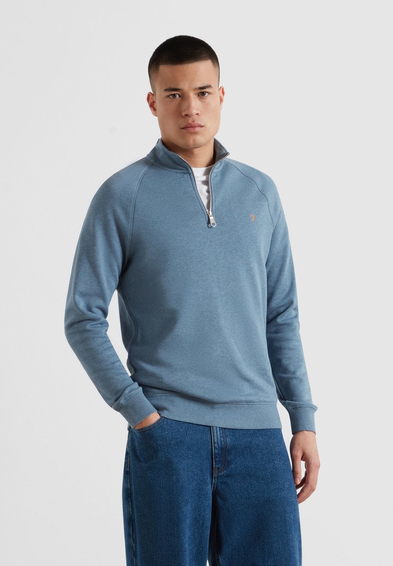 Mann trägt hellblauen Viertel-Reißverschluss-Pullover und blaue Jeans, steht mit einer Hand in der Tasche vor weißem Hintergrund.