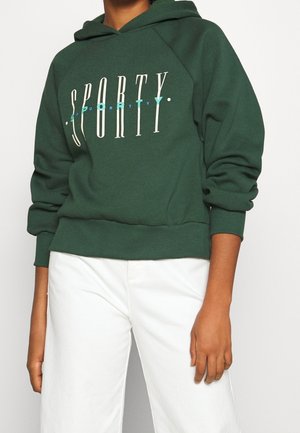 Felpa verde con una grafica frontale che recita "SPORTY" in bianco e teal. Presenta una tasca a canguro e polsini a coste. Indossata con pantaloni bianchi.