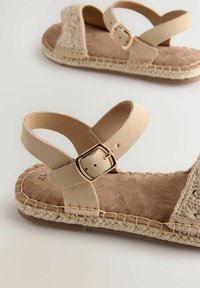 Beige sandalen met een geweven espadrille-stijl zool, voorzien van een zachte suède bovenkant en een verstelbaar enkelsluip met een gouden gespdetail.