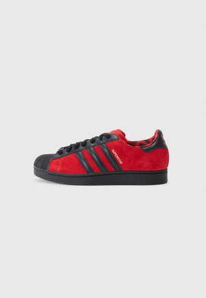 Baskets en daim rouge avec embout noir, bandes noires, lacets noirs et doublure intérieure à carreaux, vue de côté sur fond uni.