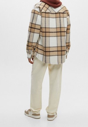 Button-up flannel shirt with beige and brown plaid pattern, worn over a maroon hooded garment. Light beige pants and sneakers complete the look.