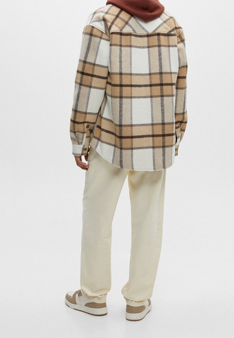Chemise en flanelle à boutons avec un motif à carreaux beige et marron, portée par-dessus un vêtement à capuche bordeaux. Un pantalon beige clair et des baskets complètent la tenue.
