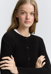 Schwarze, gerippte Strickjacke mit rundem Halsausschnitt, die drei schwarze Knöpfe an der Vorderseite und lange Ärmel hat. Der Stoff wirkt strukturiert.