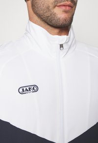 Witte zip-up jas met marineblauwe accenten, voorzien van een hoge kraag en geborduurd "JAKO SPORT" logo op de borst. Gladde, lichte stof.