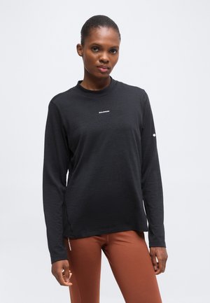 Mujer vestida con una camiseta negra de manga larga Salomon y pantalones marrones, de pie frente a un fondo liso y claro.