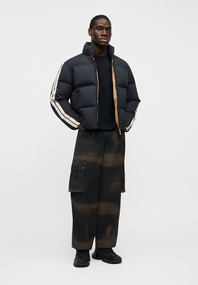 Άνδρας που φοράει μαύρο μπουφάν με μπεζ ρίγα στα μανίκια, μαύρο πουκάμισο, μαύρο και καφέ παντελόνι cargo και μαύρα αθλητικά παπούτσια, στέκεται με τα χέρια στις τσέπες.