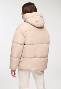 Beżowa kurtka puchowa z kapturem, charakteryzująca się pikowanym wzorem i luźnym krojem. Gładka tekstura; widoczne szwy i puchata izolacja.