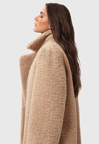 Beige, strukturierter Mantel mit hohem Kragen und weiten Ärmeln. Der Stoff wirkt flauschig und warm, was seinen gemütlichen Look unterstreicht.