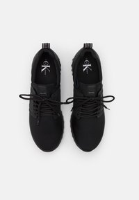 Chaussures de sport noires avec une tige en textile lisse, des lacets, et une semelle en caoutchouc présentant un motif texturé. Logo visible sur la semelle intérieure et sur le talon.