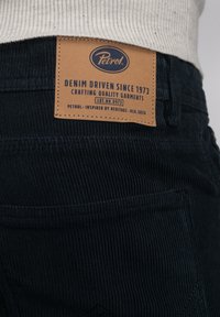 Donkerblauwe ribfluwelen broek met een tan leren label. Het label toont de merknaam "Petrol" en tekst over kwaliteit en erfgoed.