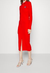 Robe rouge à manches longues avec un col, dotée d'une fermeture éclair à l'avant et d'une fente haute sur la cuisse. Assortie à des talons blancs à lanières.