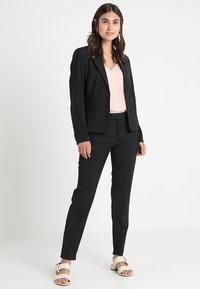 Černý oblekový blazer s dvěma předními kapsami, nošený přes světle růžový top, doplněný o černé fitted kalhoty a bílé sandály se zlatými akcenty.