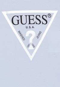 Světle modré tričko s velkým černým trojúhelníkovým logem s nápisem "GUESS U.S.A." a centrálním otazníkem, s bílými akcenty na trojúhelníku.