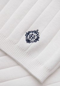 Tissu blanc côtelé avec un petit emblème floral circulaire brodé en bleu marine au centre.