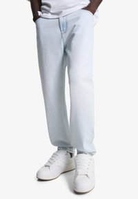 Osoba u svijetloplavim ravnim trapericama s rukama u džepovima i bijelim tenisicama na bijeloj pozadini.